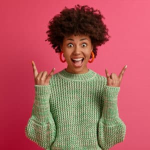 happy carefree dark skinned rebellious young woman enjoys awesome music makes rock n roll gesture has fun on music festival or cool event wears casual jumper poses against pink wall Keemya Formula of success.